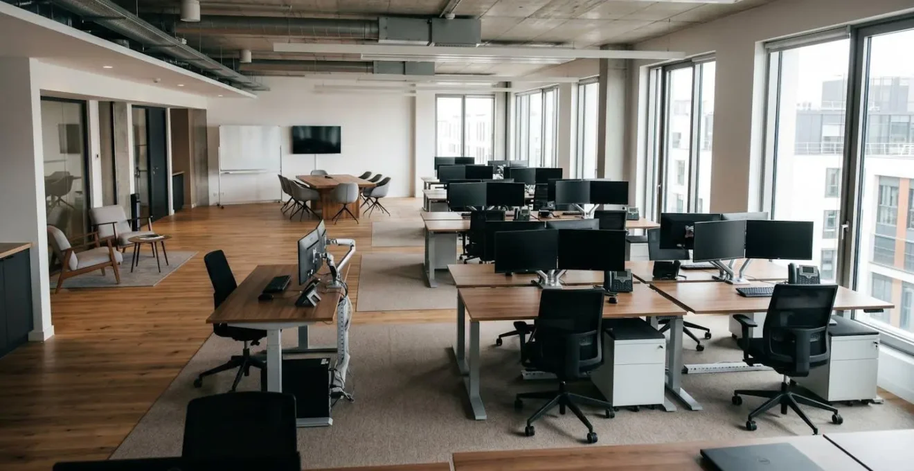 Vue d'ensemble d'un espace de bureaux professionnel moderne avec plusieurs postes de travail équipés de bureaux réglables, fauteuils ergonomiques et rangements mobiles, baigné de lumière naturelle